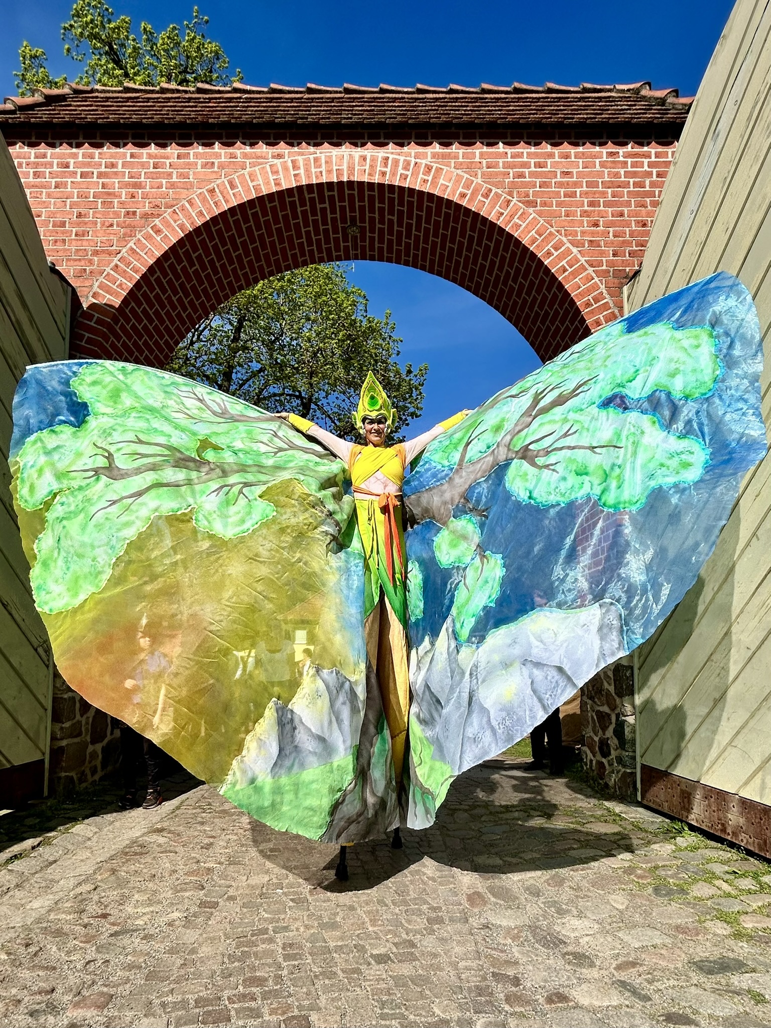 Walking Act auf Stelzen mit großen Flügeln, die einen Baum zeigen, vor einem Torbogen